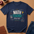 A navy blue graphic t-shirt featuring a math-themed design with equations, a lightbulb, a calculator, and the words 'MATH TEACHER' in white and teal. The shirt is laid flat on a brown cork background with lifestyle props including an apple, a coffee cup, a planner, and jeans.