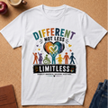 A white short-sleeve graphic T-shirt laid flat on a cork board background, featuring the phrase “DIFFERENT NOT LESS LIMITLESS” above a multicolor heart and diverse human figures. The shirt is styled with partial props including an apple, a coffee cup, a notebook, and jeans around it.