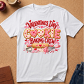 A white short-sleeve graphic T-shirt laid flat on a cork board background, featuring a Valentine's Day cookie baking theme with the words 'Valentines Day Cookie Baking Crew' and baking tools illustrated in pink, red, and gold tones.
