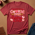 A heathered red short-sleeve t-shirt laid flat on a brown textured surface, featuring a Valentine's Day cookie-themed graphic that reads 'Valentine's Day Cookie Baking Crew' with pink hearts, striped cookies, and a rolling pin.