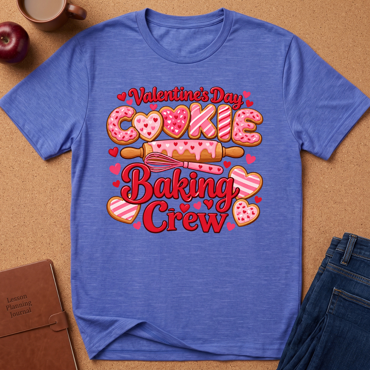 A heathered blue short-sleeve t-shirt with a colorful 'Valentine's Day Cookie Baking Crew' graphic, styled in a flat-lay on a cork background with an apple, coffee mug, notebook, and jeans.
