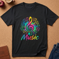 A black short-sleeve t-shirt laid flat on a cork surface, featuring a colorful graphic of a treble clef inside a music note, musical staff lines, and the word 'Music' in rainbow gradient text. Styled flat-lay props include an apple, a coffee cup, a journal, and jeans.