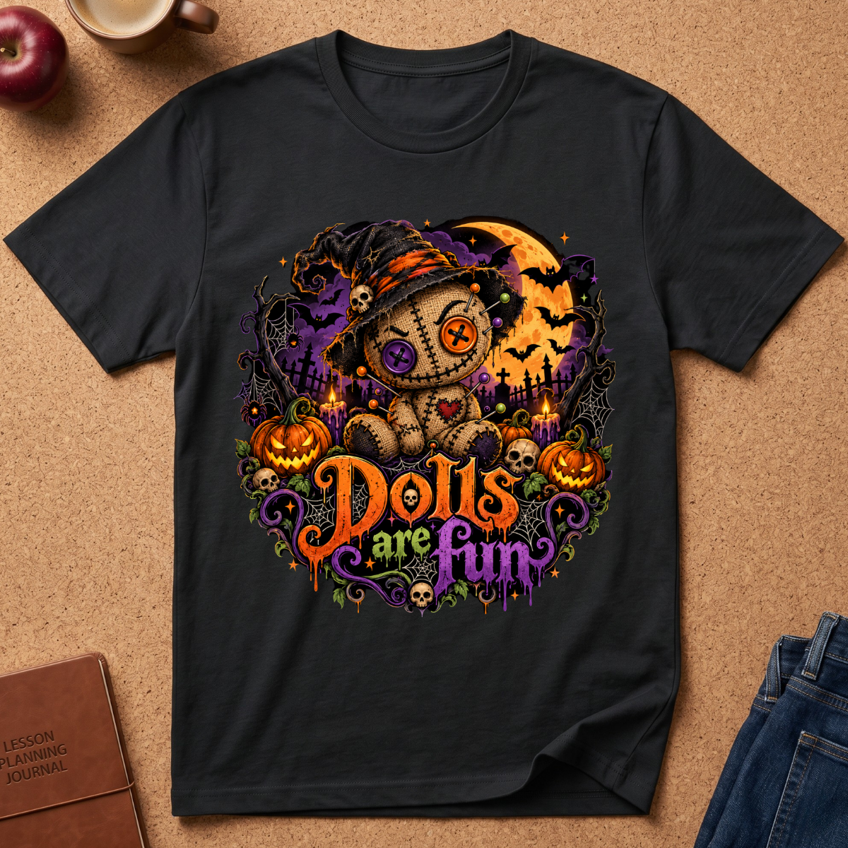 A black short-sleeve graphic T-shirt laid flat on a tan cork-like surface, featuring a Halloween-themed teddy bear illustration with pumpkins, bats, a moon, and the phrase 'Dolls are Fun' in orange and purple lettering. Partial props around the shirt include an apple, a cup, a notebook, and jeans.