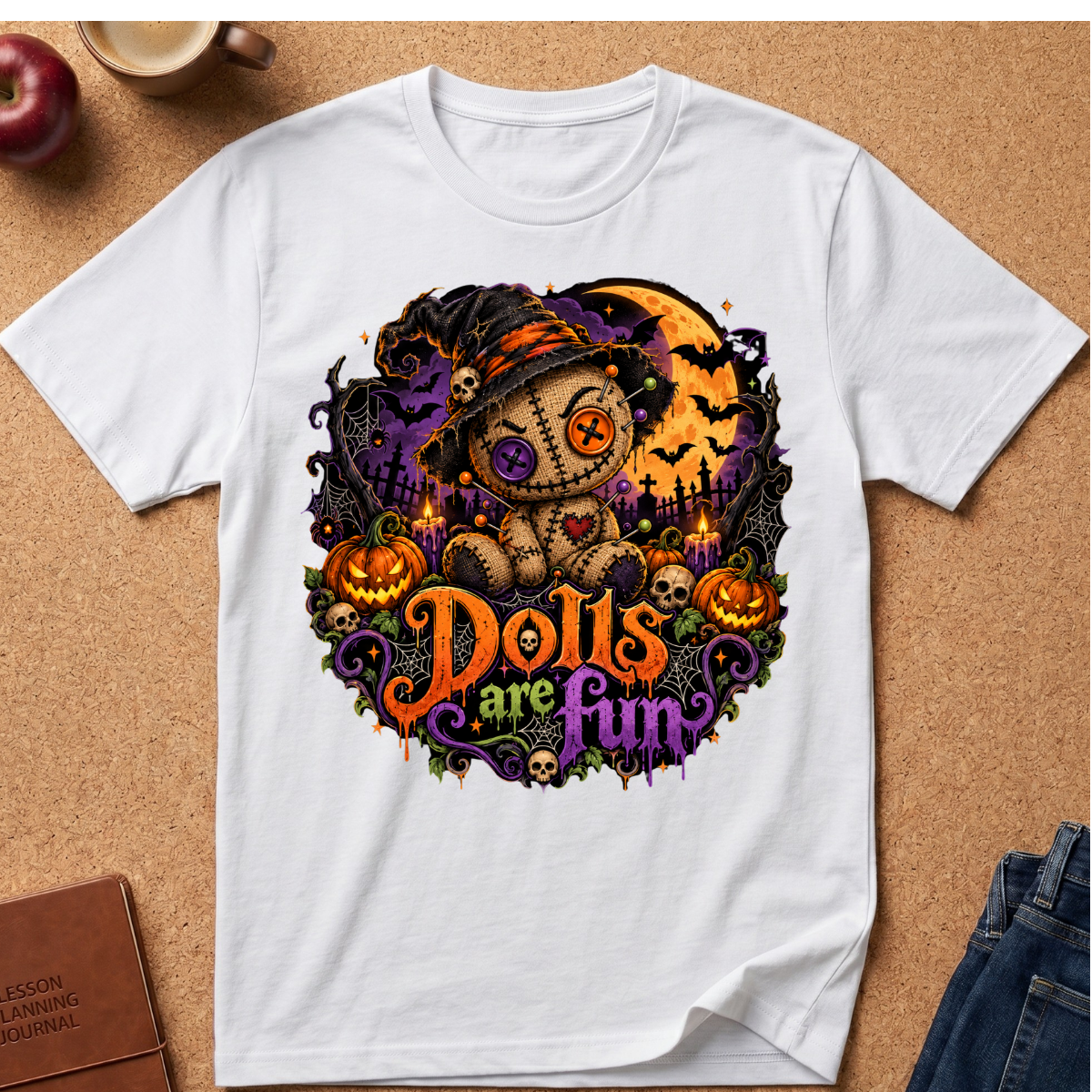 A white short-sleeve graphic T-shirt laid flat on a cork surface, featuring a Halloween-themed illustration with a doll wearing a witch hat, pumpkins, bats, a full moon, and the text 'Dolls are Fun' in orange and purple. Surrounding props include a mug, apple, notebook, and jeans.