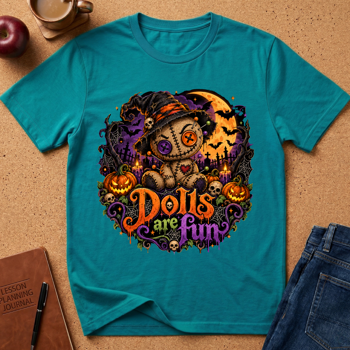 A teal short-sleeve graphic T-shirt laid flat on a cork surface, surrounded by styled props including an apple, a coffee cup, a notebook, and jeans. The shirt features a large Halloween-themed central graphic with a doll, witch hat, pumpkins, bats, a full moon, and the phrase 'Dolls are Fun' in orange and purple lettering.