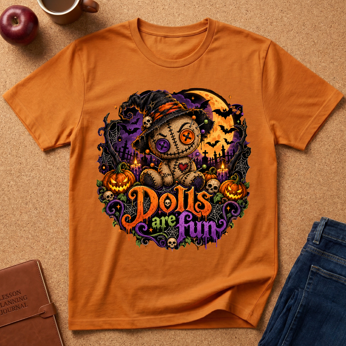 An orange short-sleeve graphic T-shirt laid flat on a cork surface, featuring a Halloween-themed illustration of a teddy bear in a witch hat with pumpkins, bats, a full moon, and the phrase 'Dolls are Fun.' Partial jeans, a notebook, a cup, and an apple are visible around it as styling props.