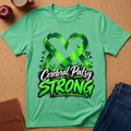 A green short-sleeve T-shirt laid flat on a cork surface, featuring a large graphic with a heart-shaped ribbon and the words 'Cerebral Palsy STRONG' in white and bright green. Props including an apple, a cup, a brown journal, and blue jeans are partially visible around the shirt.