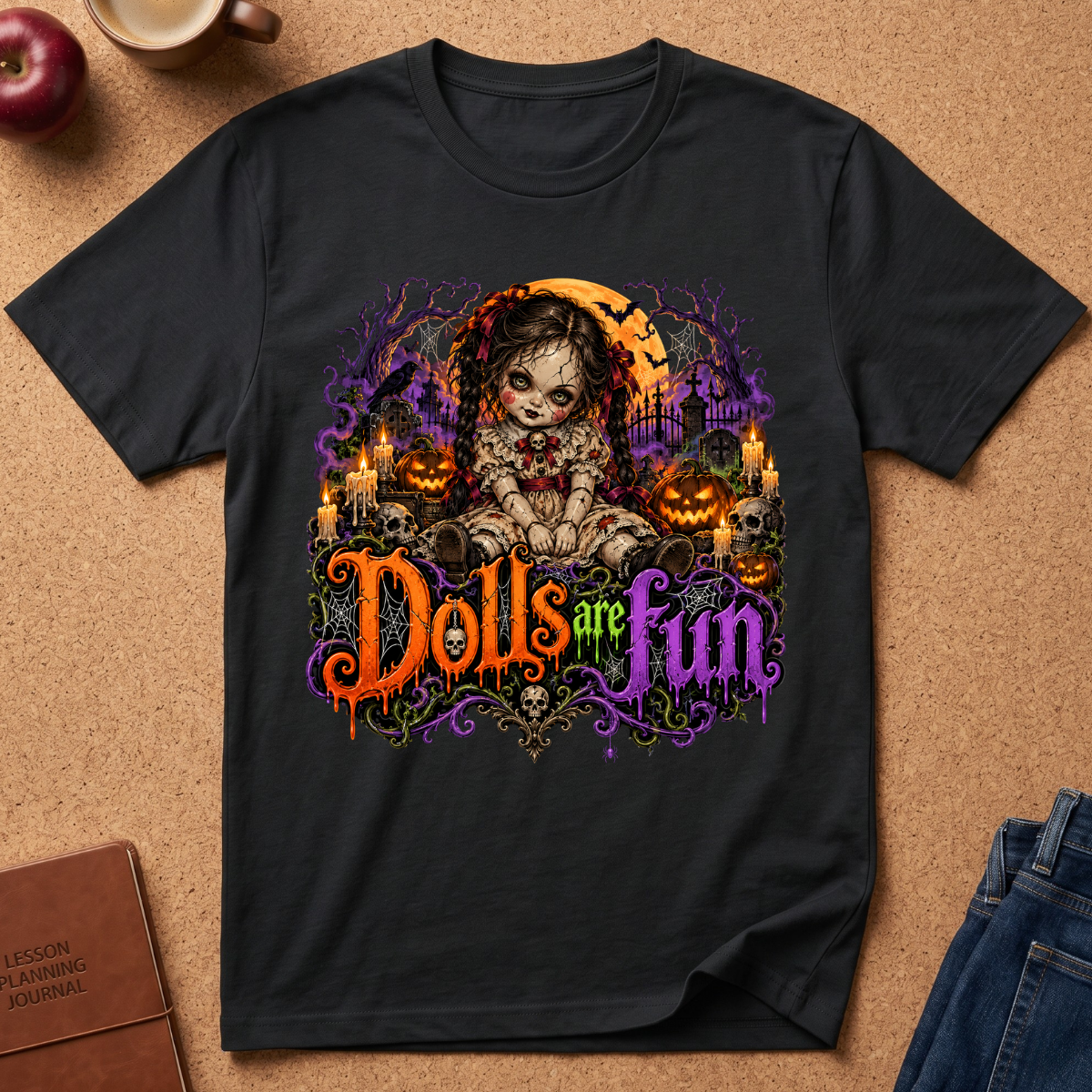 A black short-sleeve graphic T-shirt laid flat on a cork surface, featuring a Halloween-themed illustration of a doll with candles, pumpkins, a moon, and the phrase 'Dolls are fun' in orange, purple, and green lettering. Partial props including an apple, mug, journal, and jeans are visible around the shirt.