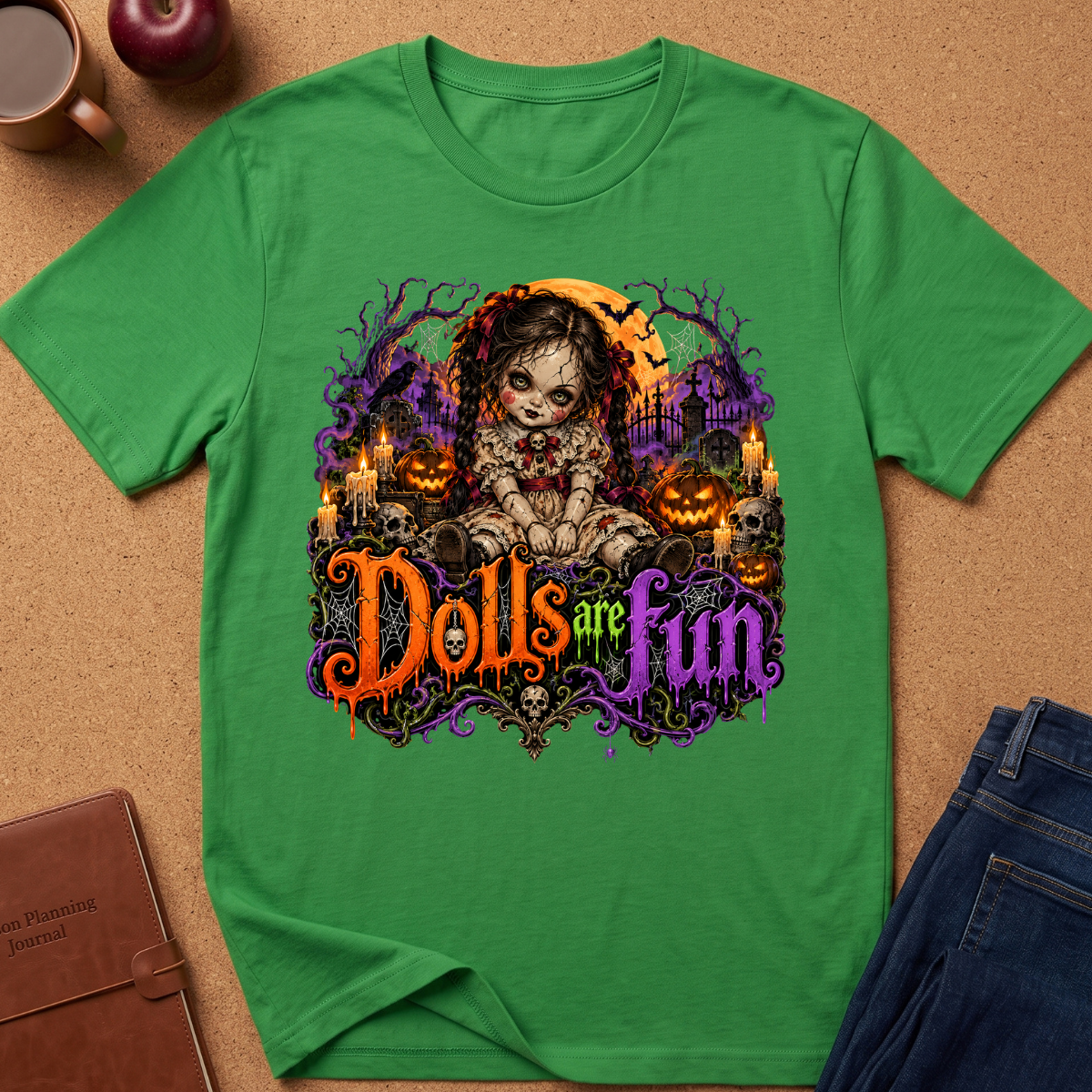 A green short-sleeve t-shirt laid flat on a cork background, featuring a colorful Halloween graphic of a creepy doll character with the text 'Dolls are fun' in orange and purple. Styled flat-lay props include a brown journal, an apple, a coffee mug, and blue jeans.