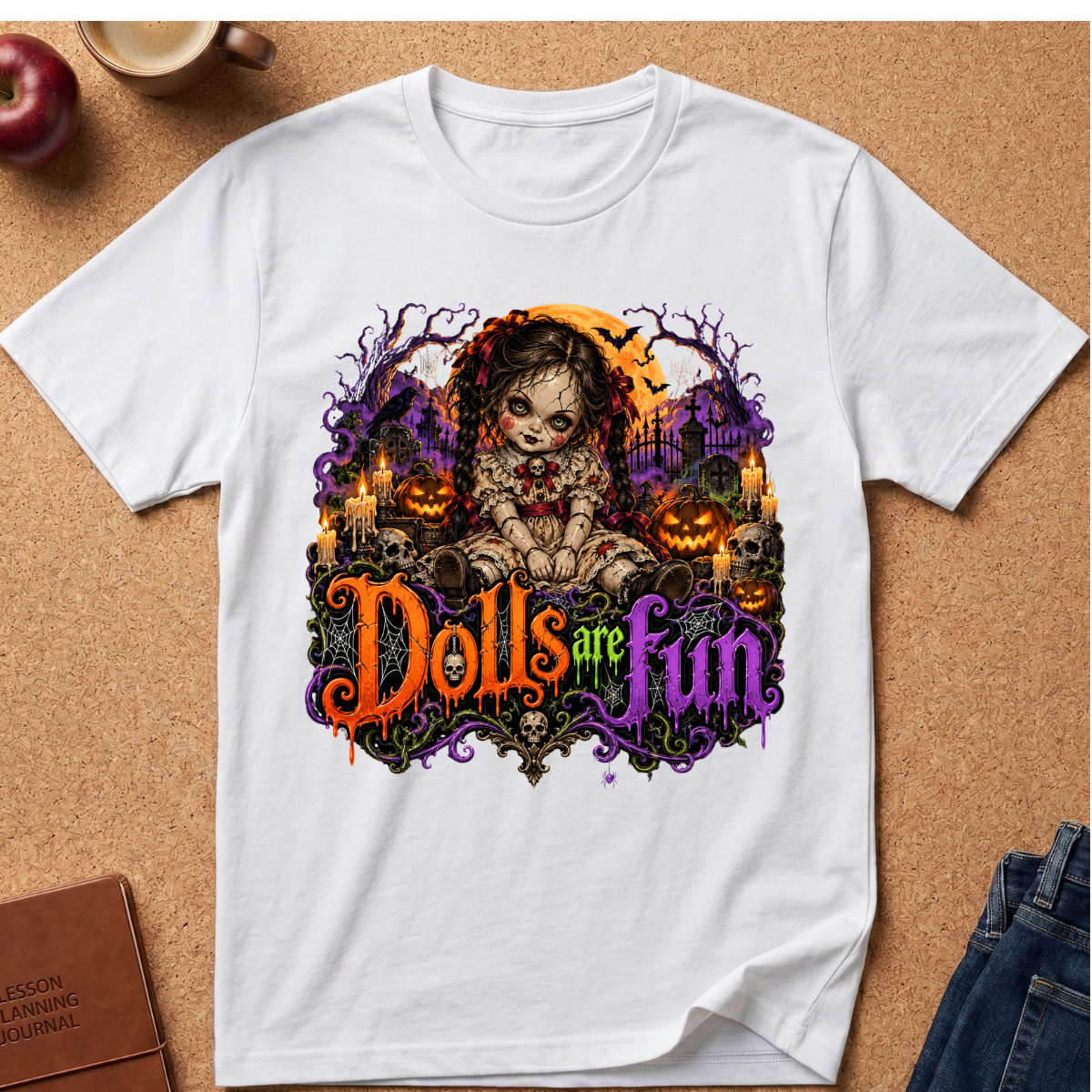 A white short-sleeve graphic T-shirt laid flat on a cork surface, featuring a Halloween-themed illustration with a doll, pumpkins, candles, and the phrase 'Dolls are fun' in orange and purple lettering. Surrounding props include a mug, apple, notebook, and jeans.