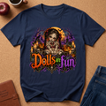 A navy blue graphic t-shirt laid flat on a brown cork board background, surrounded by lifestyle props including an apple, a coffee cup, a journal, and jeans. The t-shirt features a detailed Halloween graphic of a creepy doll character in front of a full moon and pumpkins, with the text 'Dolls are fun' in stylized orange, green, and purple lettering.
