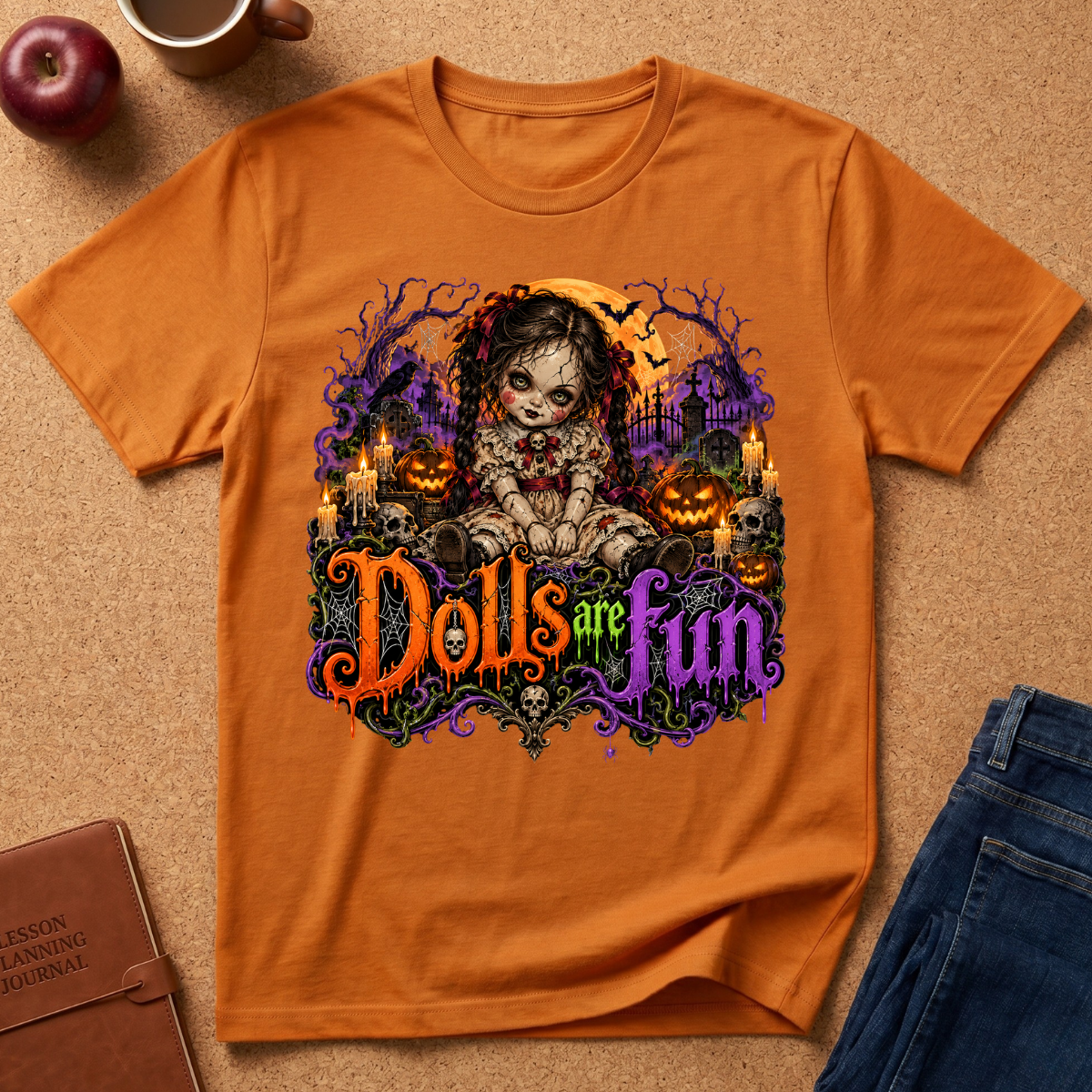 An orange short-sleeve graphic T-shirt laid flat on a cork surface, featuring a Halloween-themed illustration of a doll with candles, pumpkins, and the phrase 'Dolls are fun' in colorful lettering. Surrounding props include an apple, a mug, a notebook, and folded blue jeans.