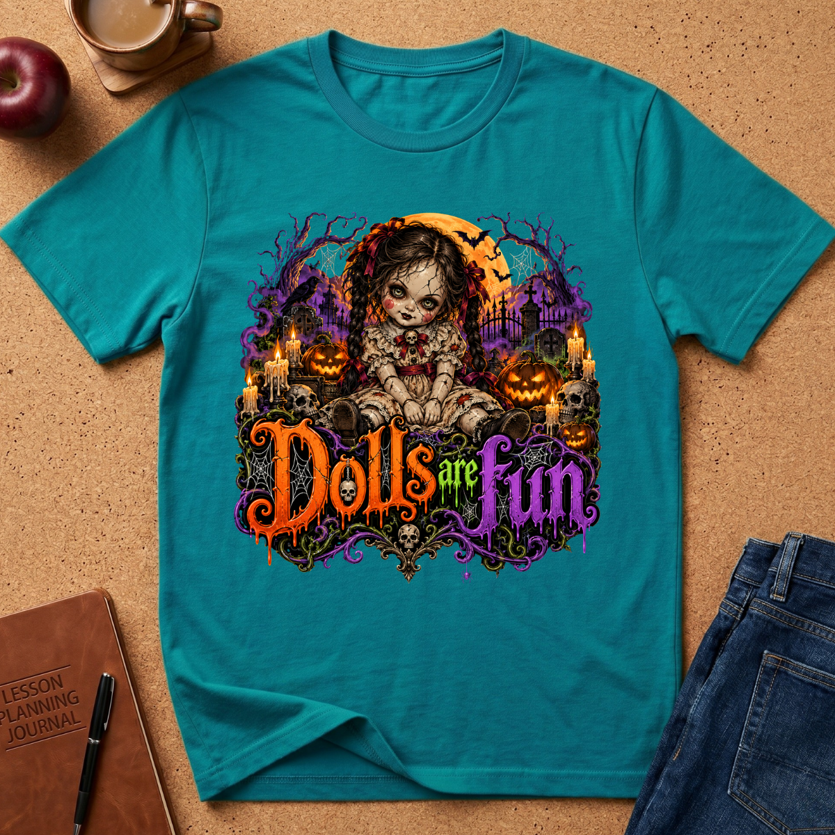 A teal short-sleeve graphic T-shirt laid flat on a cork surface, surrounded by styled props including an apple, a coffee cup, a notebook, and jeans. The shirt features a large Halloween-themed graphic with a doll-like character, candles, pumpkins, and the phrase 'Dolls are fun' in colorful lettering.