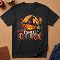 A black short-sleeve graphic T-shirt laid flat on a cork surface, featuring a Halloween design with a jack-o'-lantern, witch hat, broom, cauldron, books, stars, and the phrase “I SMELL CHILDREN” in colorful lettering.