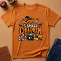 An orange short-sleeve t-shirt with a colorful Halloween graphic that reads 'I SMELL CHILDREN' in stylized lettering, featuring a jack-o'-lantern, witch hat, cauldron, books, and bats, laid flat on a cork surface with partial jeans, an apple, and a mug visible around it.