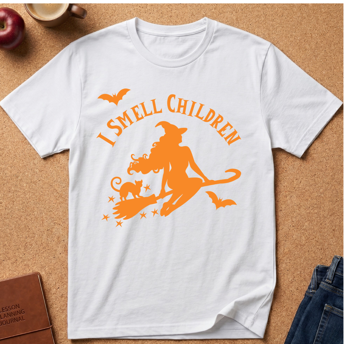 A white short-sleeve crewneck t-shirt with an orange graphic print reading 'I SMELL CHILDREN' above a witch riding a broomstick with a cat, stars, and flying bats, laid flat on a cork background with styled props.