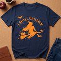 A navy blue short-sleeve t-shirt with an orange graphic of a witch riding a broomstick with a cat, accompanied by the text 'I SMELL CHILDREN' and small bats. The shirt is displayed flat on a cork background with lifestyle props including an apple, a coffee cup, a brown journal, and blue jeans.