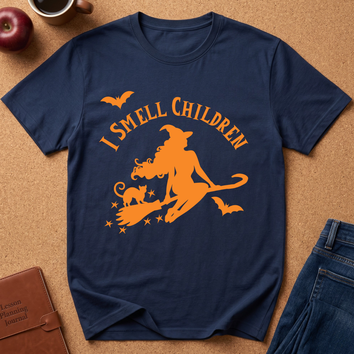 A navy blue short-sleeve t-shirt with an orange graphic of a witch riding a broomstick with a cat, accompanied by the text 'I SMELL CHILDREN' and small bats. The shirt is displayed flat on a cork background with lifestyle props including an apple, a coffee cup, a brown journal, and blue jeans.