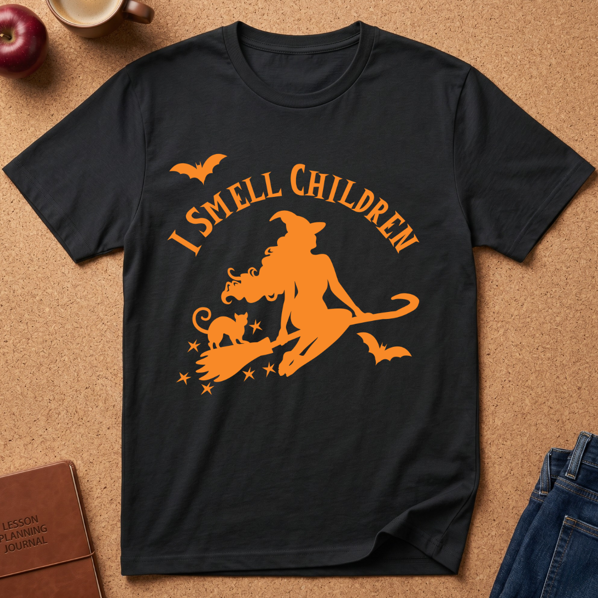 A black short-sleeve graphic T-shirt laid flat on a cork surface, featuring an orange witch riding a broomstick with a cat, stars, bats, and the text 'I SMELL CHILDREN.' Partial props around the shirt include an apple, a mug, a journal, and jeans.