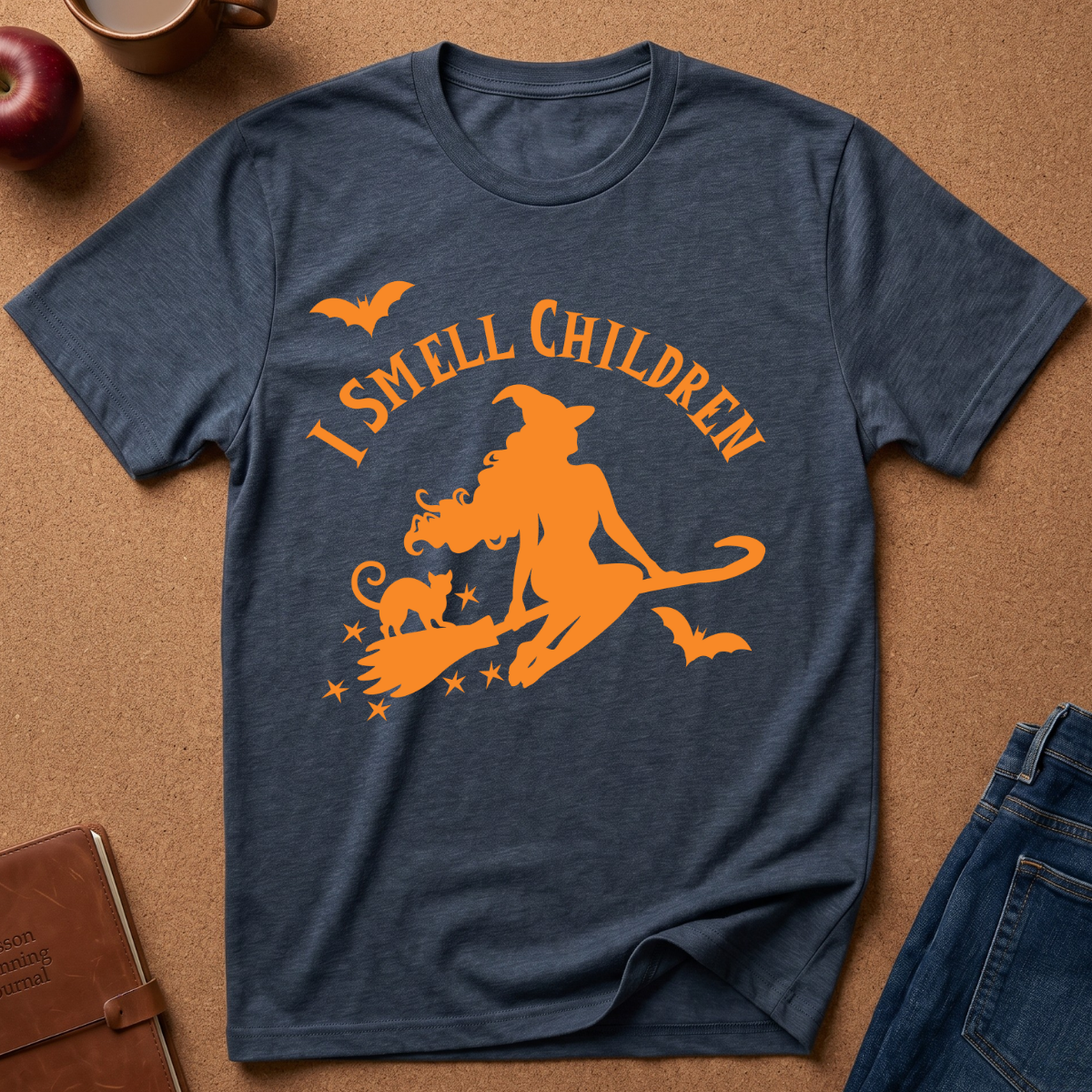 A heather navy blue short-sleeve t-shirt with a bright orange graphic of a witch riding a broomstick with a cat, accompanied by the text 'I SMELL CHILDREN' and small bats, laid flat on a cork background with jeans, a notebook, and an apple partially visible.