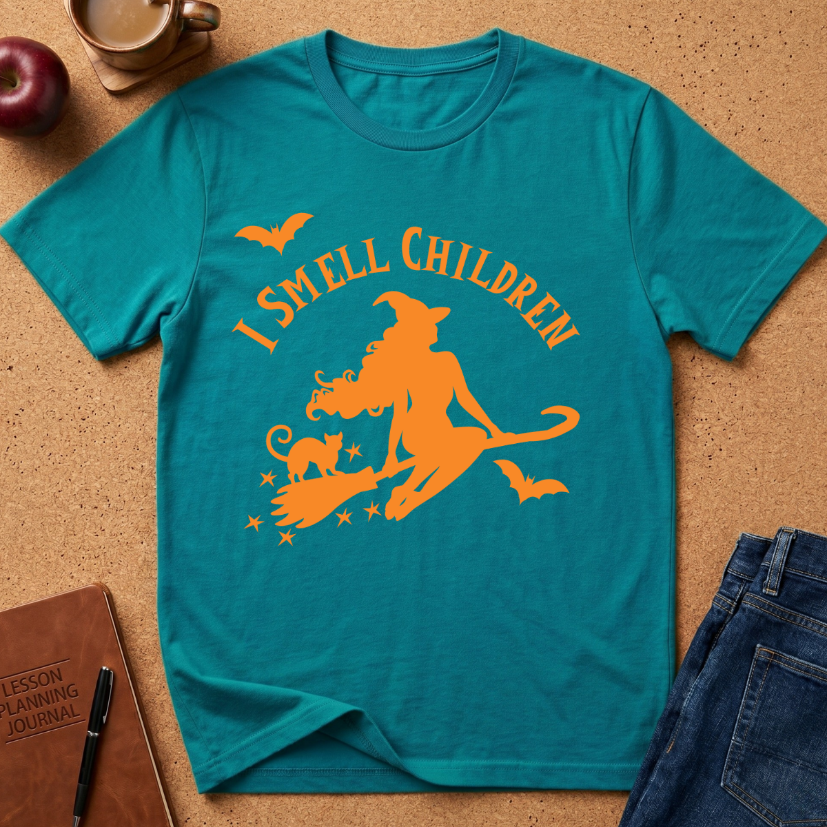 A teal short-sleeve graphic T-shirt laid flat on a cork surface, featuring an orange witch riding a broomstick with a cat and the text 'I SMELL CHILDREN' above. Surrounding props include a coffee cup, apple, notebook, pen, and jeans.