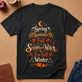 A black short-sleeve graphic T-shirt laid flat on a cork surface, featuring the phrase 'Spring Summer Fall Season of the Witch 2nd Fall Winter' in decorative orange, yellow, green, and white lettering with autumn leaves, sunflowers, a witch hat, and pumpkins. Partial props including an apple, a mug, a journal, and jeans are visible around the shirt.