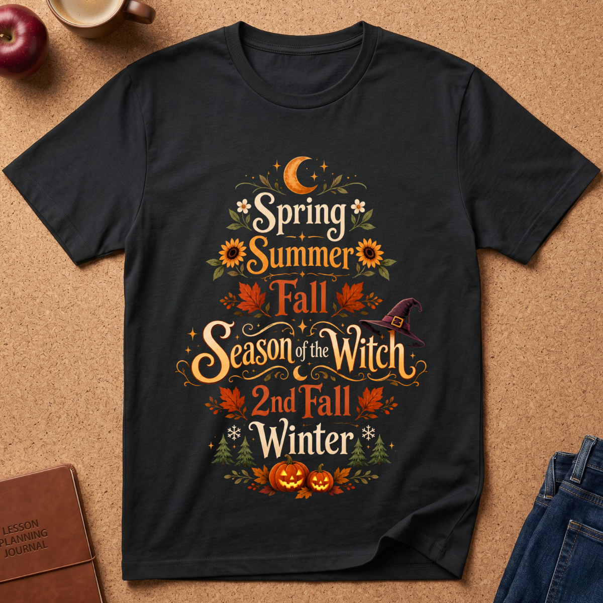 A black short-sleeve graphic T-shirt laid flat on a cork surface, featuring the phrase 'Spring Summer Fall Season of the Witch 2nd Fall Winter' in decorative orange, yellow, green, and white lettering with autumn leaves, sunflowers, a witch hat, and pumpkins. Partial props including an apple, a mug, a journal, and jeans are visible around the shirt.