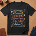 A black graphic t-shirt laid flat on a cork background, featuring a colorful seasonal design that reads 'Spring Summer Fall Season of Witch 2nd Fall Winter' with Halloween-themed illustrations including pumpkins, ghosts, sunflowers, and leaves.