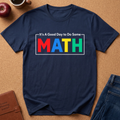 A navy blue short-sleeve t-shirt with a colorful graphic reading 'It's A Good Day to Do Some MATH' in red, blue, yellow, and green letters, styled in a flat lay on a brown cork background with an apple, coffee cup, and jeans nearby.
