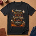 A black graphic t-shirt laid flat on a cork background, featuring a colorful autumn-themed design with the text 'Spring, Summer, Fall, Season of the Witch, 2nd Fall, Winter' surrounded by pumpkins, ghosts, a witch hat, and leaves. Styled flat-lay props include an apple, a coffee cup, a journal, and jeans.