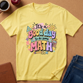 A yellow short-sleeve graphic T-shirt laid flat on a cork surface, surrounded by lifestyle props including a coffee mug, apple, notebook, and jeans. The shirt features a colorful retro-style math-themed graphic that reads 'It's a good day to do some MATH' with doodles of a smiley face, calculator, pencil, and geometric shapes.