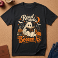 A black graphic t-shirt laid flat on a cork background, featuring a Halloween-themed design with a ghost reading a book, pumpkins, bats, stars, and the text 'Read More Booooo-ks'.