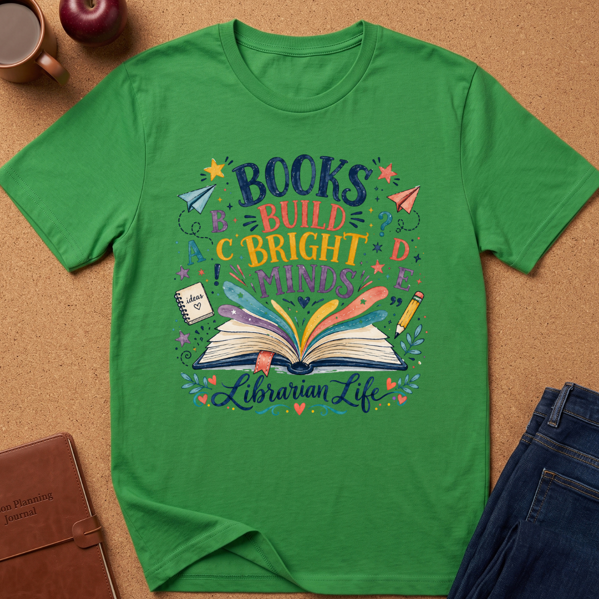 Irish Green short-sleeve graphic T-shirt laid flat on a cork surface, surrounded by lifestyle props including a coffee cup, apple, notebook, and jeans. The shirt features a colorful printed design with the words 'BOOKS BUILD BRIGHT MINDS' and 'Librarian Life' around an open book illustration.
