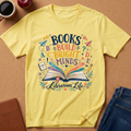 A yellow short-sleeve graphic T-shirt laid flat on a cork surface, surrounded by lifestyle props including a coffee cup, apple, notebook, and jeans. The shirt features a colorful printed design with the words 'BOOKS BUILD BRIGHT MINDS' and 'Librarian Life' around an open book illustration.