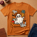 An orange short-sleeve graphic T-shirt laid flat on a cork surface, surrounded by styled props including an apple, a mug, a notebook, and folded blue jeans. The shirt features a Halloween-themed cartoon ghost with books, an apple, bats, stars, and the text 'You Are All Booooooful'.