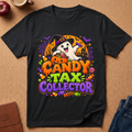 A black short-sleeve graphic T-shirt laid flat on a cork surface, featuring a colorful Halloween design with a ghost, pumpkin, bats, candy corn, and the text 'CANDY TAX COLLECTOR.' Partial props around the shirt include an apple, a cup, a journal, and jeans.
