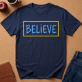 Navy Blue Graphic tee wth the text "Believe" in a blue hand drawn font inside a Yellow Rectangle.  