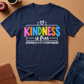 A navy blue graphic t-shirt laid flat on a brown cork background, featuring the text 'KINDNESS is free SPRINKLE IT EVERYWHERE' in colorful and white lettering. The shirt is styled with a brown leather journal, an apple, a coffee mug, and blue jeans.