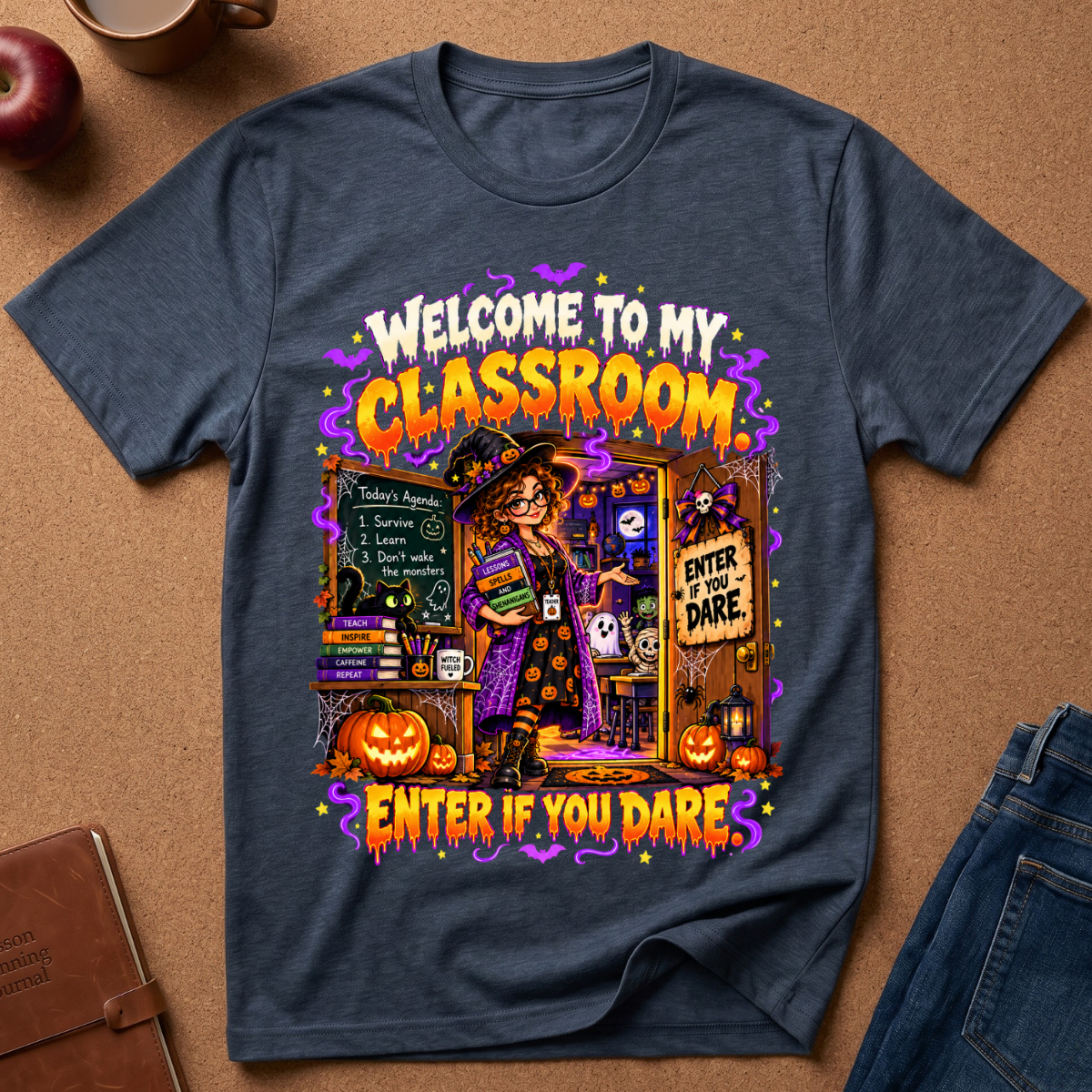 A navy blue graphic t-shirt laid flat on a brown cork background, featuring a colorful Halloween-themed design with the text 'WELCOME TO MY CLASSROOM' and 'ENTER IF YOU DARE'.