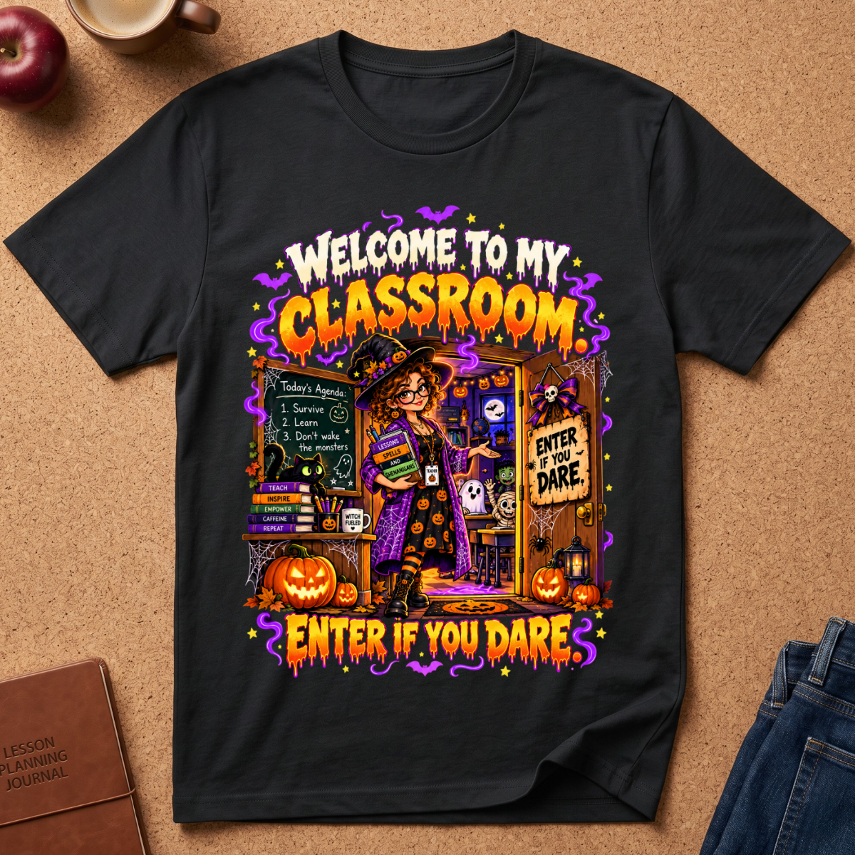 A black graphic t-shirt laid flat on a cork background, featuring a colorful Halloween-themed design with the text 'WELCOME TO MY CLASSROOM' and 'ENTER IF YOU DARE.' A witch, pumpkins, bats, and spooky decorations are illustrated on the front.