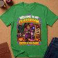 A green short-sleeve graphic T-shirt laid flat on a cork surface, surrounded by styled props including a brown notebook, an apple, a mug, and blue jeans. The shirt features a large Halloween-themed classroom graphic with the text 'WELCOME TO MY CLASSROOM' and 'ENTER IF YOU DARE.