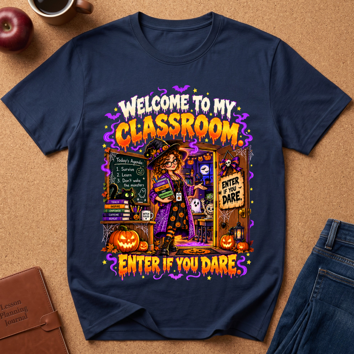 A navy blue graphic t-shirt laid flat on a brown cork board background, surrounded by lifestyle props including a brown leather journal, a red apple, a coffee mug, and blue jeans. The t-shirt features a colorful Halloween-themed graphic with the text 'WELCOME TO MY CLASSROOM' and 'ENTER IF YOU DARE' in orange and yellow, depicting a witch, pumpkins, and a chalkboard.