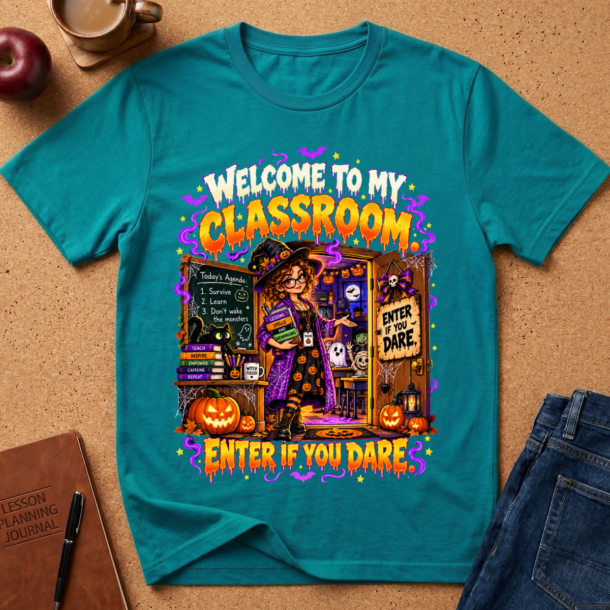 A teal short-sleeve graphic T-shirt laid flat on a cork surface, surrounded by styled props including an apple, a notebook, a pen, and jeans. The shirt features a large Halloween-themed classroom illustration with the text 'WELCOME TO MY CLASSROOM' and 'ENTER IF YOU DARE.
