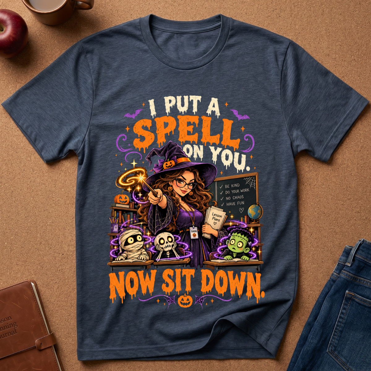 A navy blue graphic t-shirt laid flat on a brown cork background, featuring a Halloween-themed illustration of a witch casting a spell with the text 'I PUT A SPELL ON YOU. NOW SIT DOWN.
