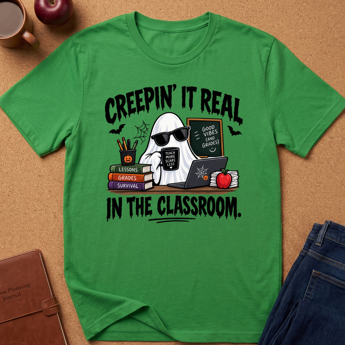 A green short-sleeve graphic T-shirt laid flat on a cork surface, surrounded by styled props including a brown journal, an apple, a mug, and blue jeans. The shirt features a Halloween-themed classroom graphic with the text 'CREEPIN' IT REAL IN THE CLASSROOM' and an illustration of a ghost in sunglasses holding a laptop, with books, bats, and an apple.