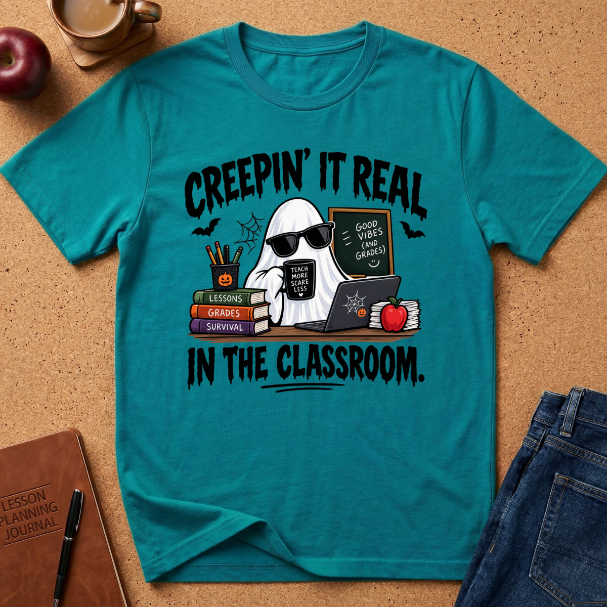 A tropical blue graphic t-shirt laid flat on a cork background, featuring a ghost character wearing sunglasses and holding a laptop next to books, an apple, and a chalkboard. The text on the shirt reads 'CREEPIN' IT REAL IN THE CLASSROOM.