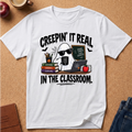 A white short-sleeve graphic T-shirt laid flat on a cork background, featuring a cartoon ghost wearing sunglasses and holding a mug that reads 'I'M NOT SLEEPING', surrounded by books, a laptop, bats, and the text 'CREEPIN' IT REAL IN THE CLASSROOM.'.