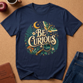 A navy blue graphic t-shirt laid flat on a cork background, featuring a whimsical 'Be Curious' design with a butterfly, magnifying glass, moon, and mushrooms. Styled flat-lay props include an apple, a coffee cup, a brown journal, and blue jeans.