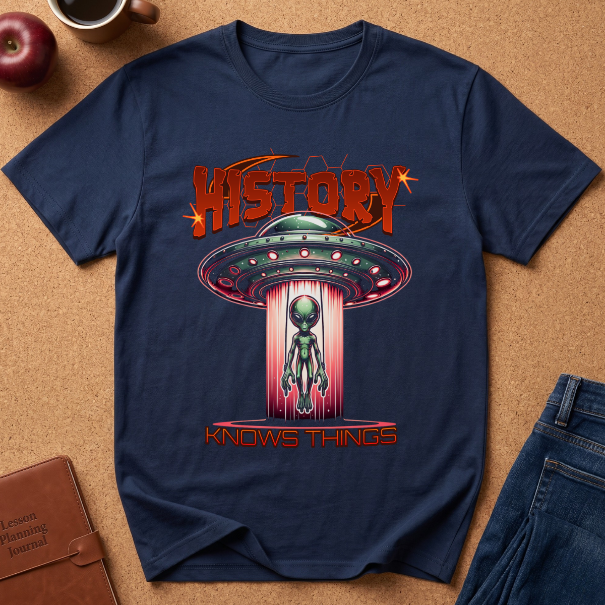 A navy blue graphic t-shirt laid flat on a cork background, featuring a retro-style alien design with the text 'HISTORY KNOWS THINGS'. The shirt is styled with a brown leather journal, a red apple, and blue jeans.