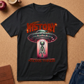 A black graphic t-shirt laid flat on a cork background, featuring a retro sci-fi design with a UFO, alien, and the text 'HISTORY KNOWS THINGS' in red and orange.